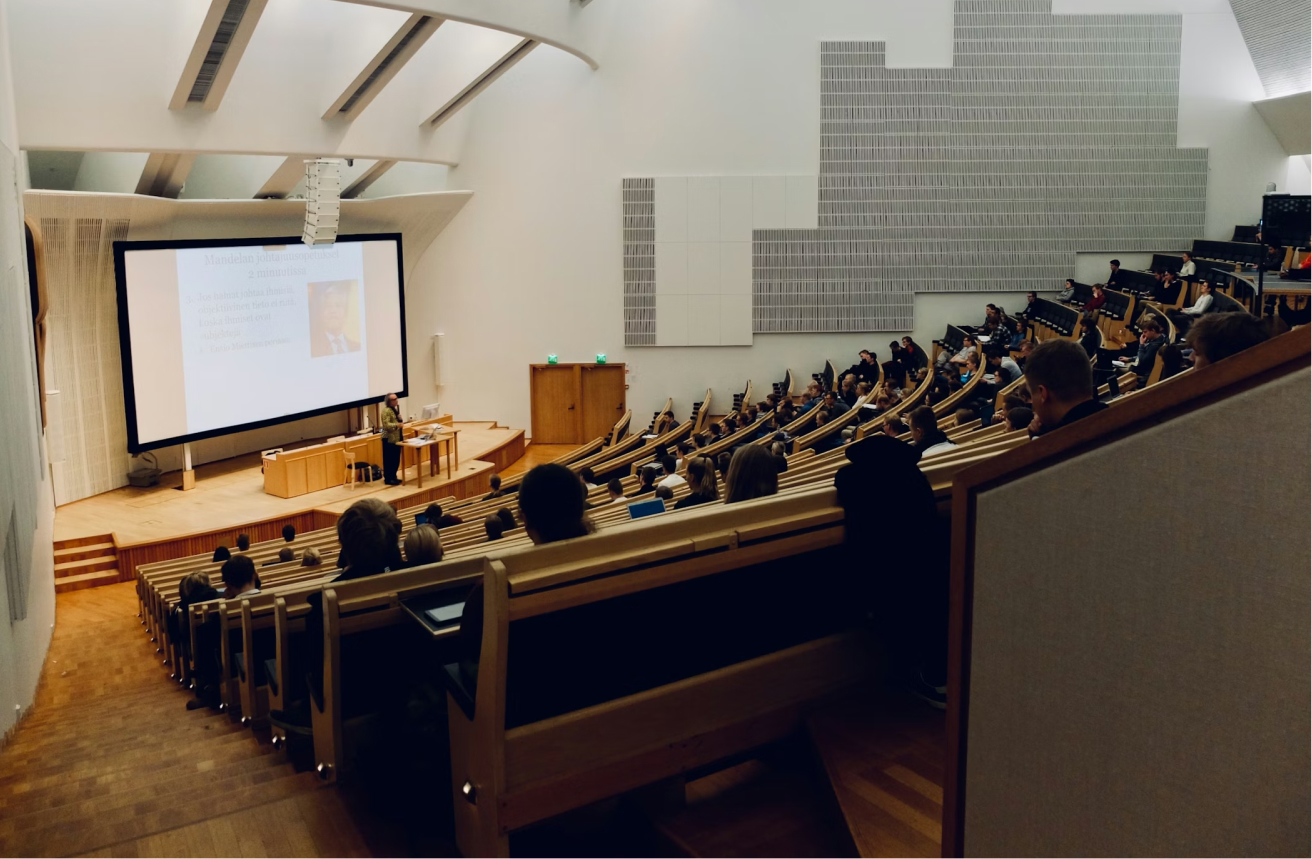 A large lecture hall full of students slopes westerly toward the smart board at the front of the room. The professor is in the middle of a lecture.