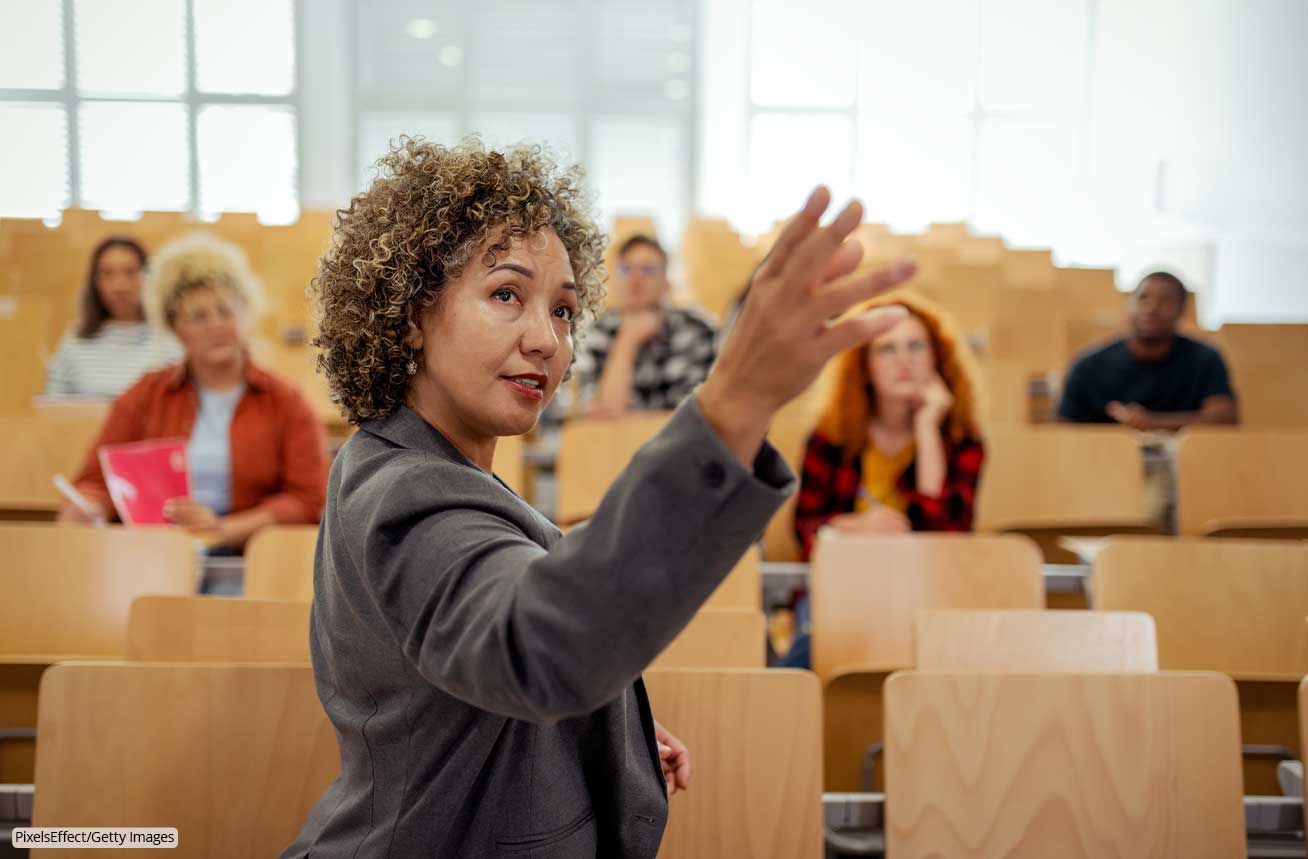 An educator gestures toward their teaching material while speaking to an engaged but small classroom of students.