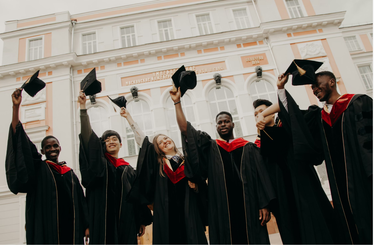 Six new graduates in black gowns and red sashes toss their caps to the sky.